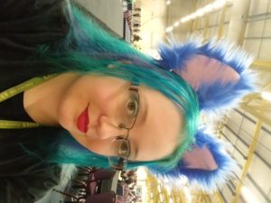 Dragon is looking into the camera with her face turned partially to the side. She wearing black clothes with a lanyard and red lipstick. Turquoise and mid blue hair is topped with royal blue fluffy ears. In the background is a convention hall