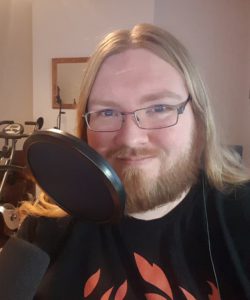 AJ is facing and smiling at the camera, a pop filter for a microphone covers part of his beard. His long blond hair is down and he is wearing a black tee-shirt