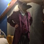 A man standing in a room wearing a black top hat and a purple waistcoat/coat tail jacket combo. His face is covered by a plague doctor mask