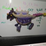 A whiteboard with numbers written on it. Attached by a magnet is a purple toy dragon with wings, head, tail and legs attached by springs
