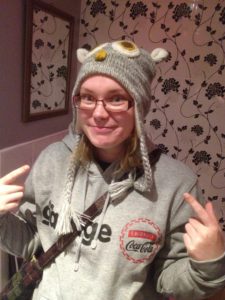 A woman standing in a grey hoodie and a grey knitted character hat, smiling and pointing to the hat. Blonde hair is visible under the hat and she wears dark red rimmed glasses